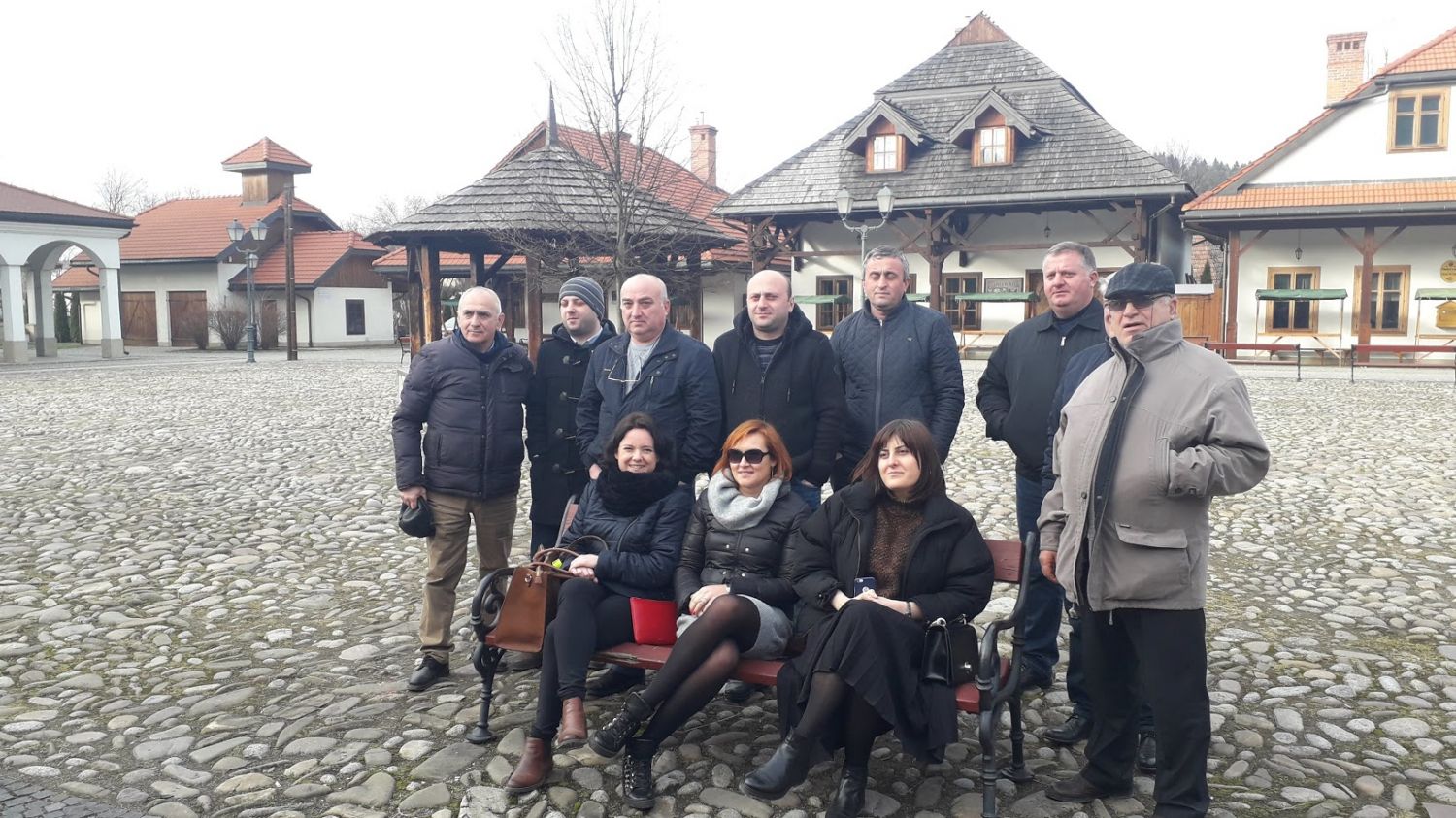 Na zdjęciu widać grupę osób pozujących do zdjęcia, część stoi, część siedzi na ławeczce. W tle widać budynki z Miasteczka Galicyjskiego, osoby stoją na brukowanym rynku.